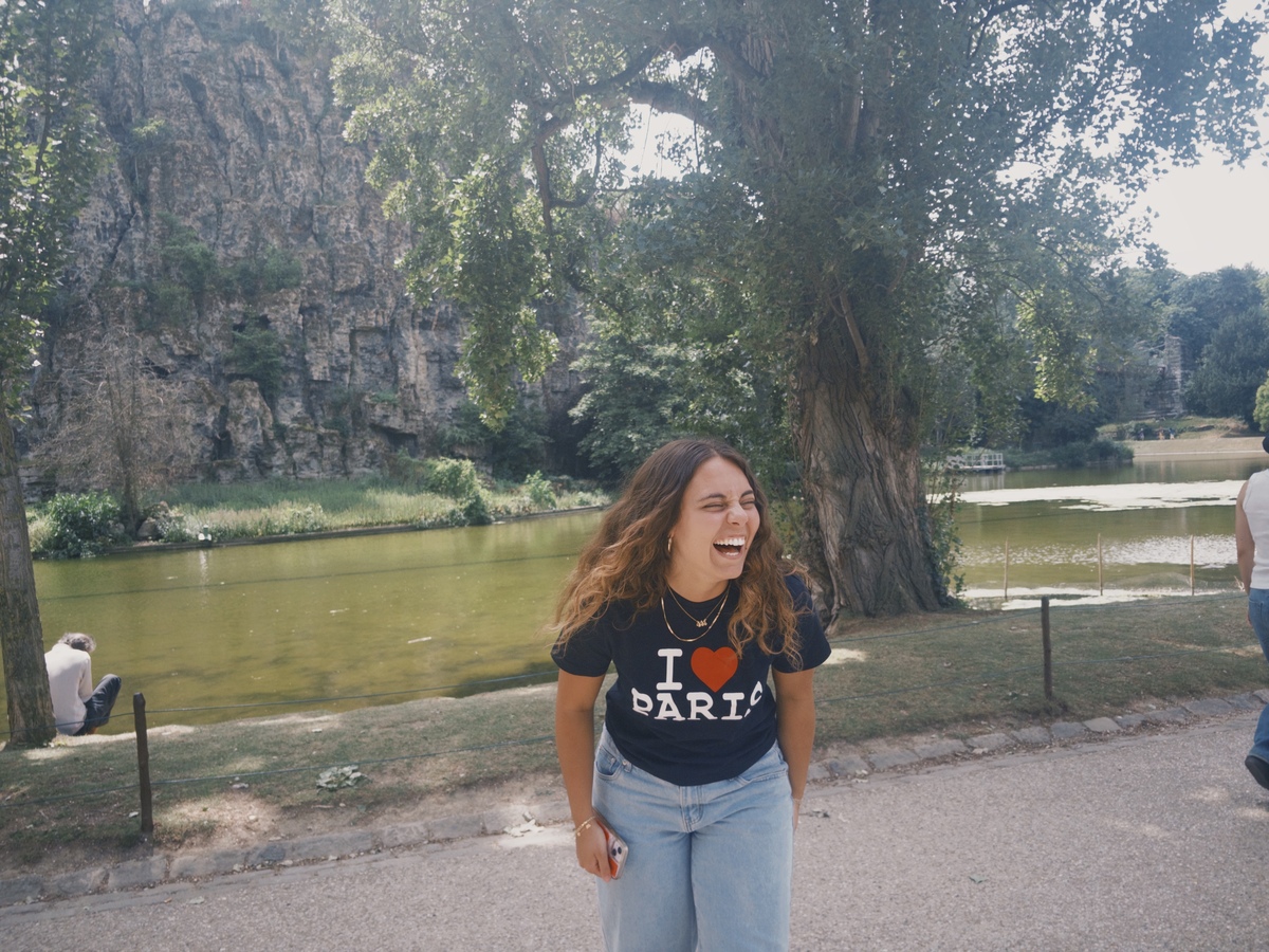 A student laughing in front of a lake in Paris, wearing an I Love Paris t-shirt